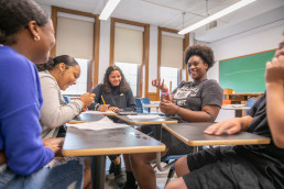 Adults sitting at desks in a circle, smiling and gesturing to each other