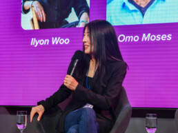 A smiling woman sits in a chair, holds a microphone, and smiles at the audience. =